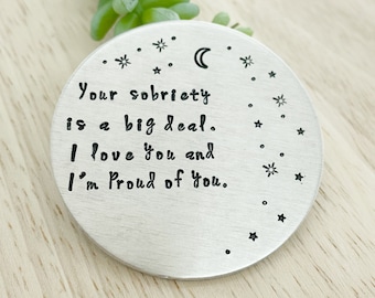 Peut inclure: Un charme rond en argent avec un message qui dit "Votre sobriété est un grand pas. Je t'aime et je suis fier de toi." Le charme est entouré d'une bordure de petites étoiles et d'un croissant de lune.