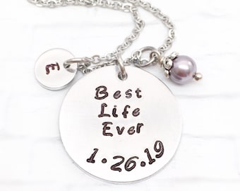 Könnte beinhalten: Silberne Halskette mit einem runden Anhänger, auf dem "Best Life Ever 1.26.19" steht, und einem kleineren Anhänger mit dem Buchstaben "E". Ein kleiner Perlenanhänger hängt an der Halskette.