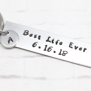 May include: Silver keychain with a small round charm with the letter "A" and a rectangular tag that reads "Best Life Ever 6.16.18"