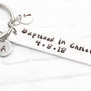May include: Silver keychain with a rectangular tag that reads "Baptized in Christ 4.8.18". The keychain also has a small charm with the letter "A" and a cross charm.