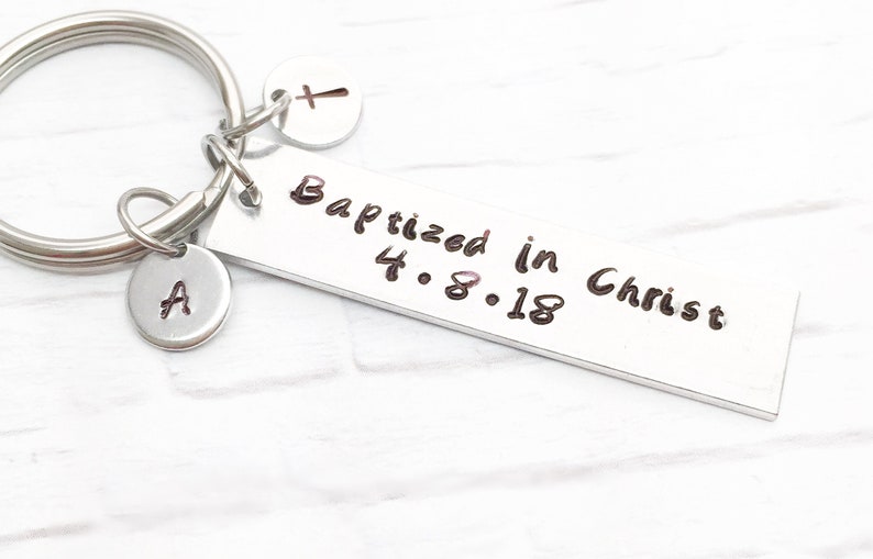May include: Silver keychain with a rectangular tag that reads "Baptized in Christ 4.8.18". The keychain also has a small charm with the letter "A" and a cross charm.