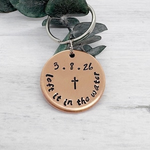 Op de afbeelding: Een koperkleurige ronde sleutelhanger met de tekst "3.8.26" boven een kruissymbool en de woorden "left it in the water" rond het kruis. De sleutelhanger is bevestigd aan een zilveren ring en rust op een wit oppervlak met groene bladeren.