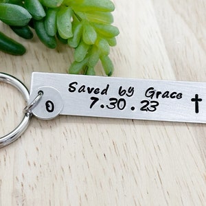 May include: Silver keychain with a rectangular tag that reads "Saved by Grace 7.30.23" and a small round charm with the letter "O" on it.
