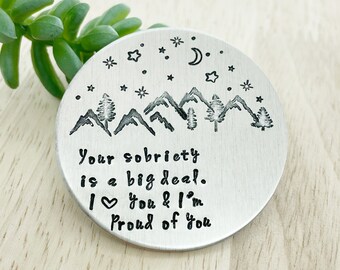 Peut inclure: Un disque en métal argenté avec un motif de chaîne de montagnes et des étoiles. Le texte "Your sobriety is a big deal. I love you & I'm proud of you" est gravé sur le disque.