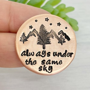 May include: A round, copper-colored metal pendant with a mountain and tree design. The pendant has the phrase "always under the same sky" engraved in black. Stars are etched around the top edge of the pendant.