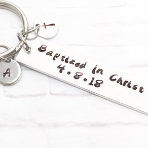 May include: Silver keychain with a rectangular tag that reads "Baptized in Christ 4.8.18". The keychain also has a small silver charm with the letter "A" and a small silver heart charm with a cross.