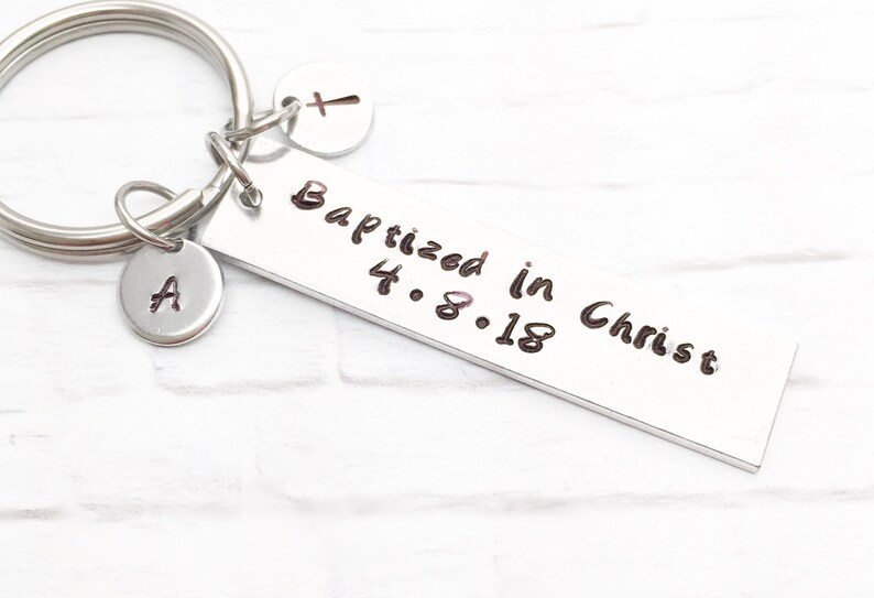 May include: Silver keychain with a rectangular tag that reads "Baptized in Christ 4.8.18". The keychain also has a small silver charm with the letter "A" and a small silver heart charm with a cross.