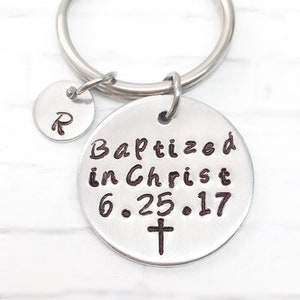 May include: A silver-colored keychain with two circular charms. The larger charm has the words "Baptized in Christ 6.25.17" and a cross. The smaller charm has the letter "R".