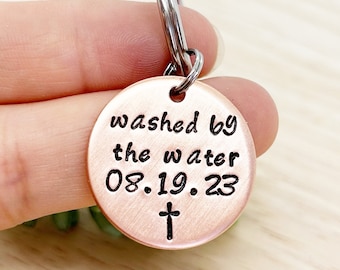 May include: A copper-coloured keychain with a round tag that reads "washed by the water 08.19.23" with a small cross below the date.