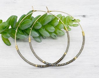 May include: A pair of gold-tone hoop earrings. The hoops are adorned with small, gray beads and gold accents. The earrings are displayed against a backdrop of green foliage and a white wooden surface.