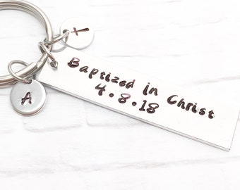 May include: Silver keyring with a rectangular tag that reads "Baptized in Christ 4.8.18". The keyring also has a small charm with the letter "A" and a cross charm.