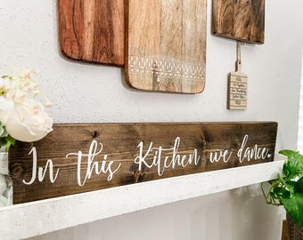 May include: A rustic wooden sign with the text "In this kitchen we dance." The sign is mounted on a white shelf with a plant in a pot on the right side of the image.