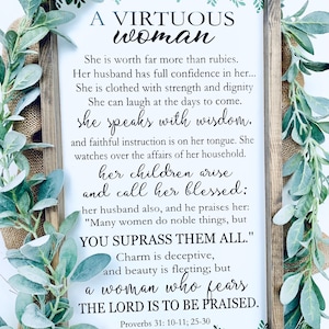 May include: A rectangular wooden sign with a dark brown frame featuring the text A VIRTUOUS WOMAN and verses from Proverbs 31: 10-11; 25-30 printed in black on a white background, accented with green leaf illustrations at the top and bottom.