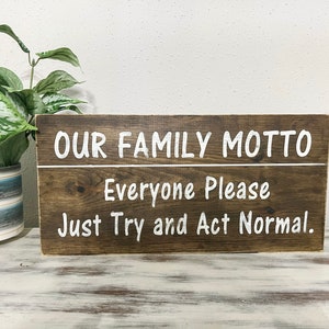 May include: A brown wooden sign with white text that reads "Our Family Motto: Everyone Please Just Try and Act Normal."