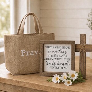 May include: A tan corduroy tote bag with the word Pray printed in white, alongside a square wooden sign featuring the text Those who leave everything in Gods hands will eventually see Gods hands in everything, and a wooden cross.