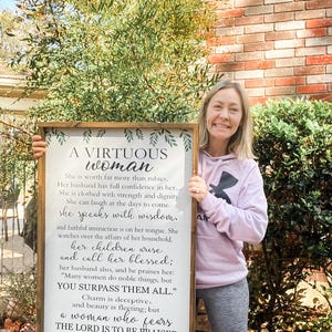 May include: A rectangular wooden framed sign featuring black printed text on a white background. The text reads A VIRTUOUS WOMAN and includes verses from Proverbs 31: 10-11, 25-30. Green leaf illustrations decorate the top and bottom borders.