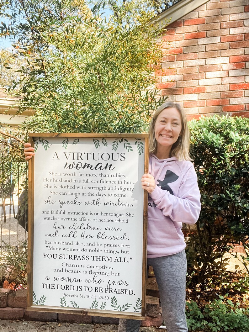May include: A rectangular wooden framed sign featuring black printed text on a white background. The text reads A VIRTUOUS WOMAN and includes verses from Proverbs 31: 10-11, 25-30. Green leaf illustrations decorate the top and bottom borders.