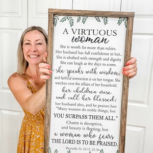 May include: A rectangular wooden sign with a dark brown frame and a white background featuring green leaf illustrations at the top and bottom. The sign displays the text A VIRTUOUS WOMAN followed by the full Proverbs 31: 10-11; 25-30 passage.