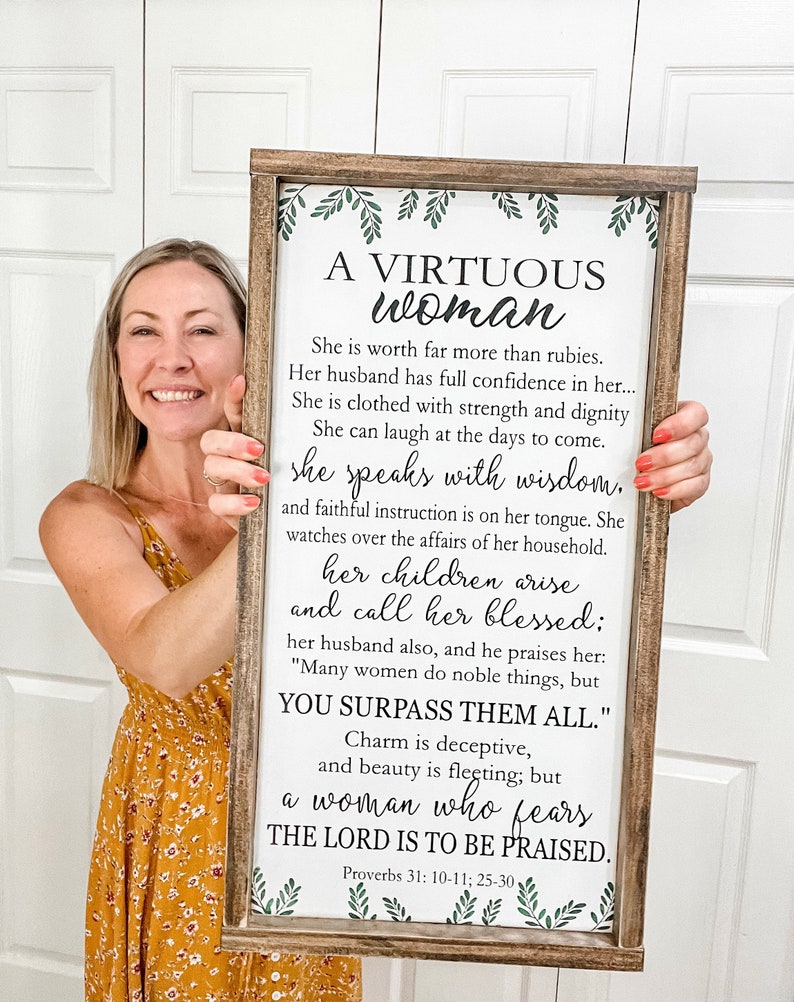 May include: A rectangular wooden sign with a dark brown frame and a white background featuring green leaf illustrations at the top and bottom. The sign displays the text A VIRTUOUS WOMAN followed by the full Proverbs 31: 10-11; 25-30 passage.