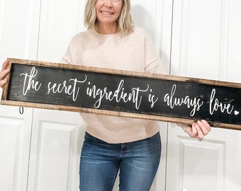 May include: A rectangular wooden sign with a black background and white cursive text that reads "the secret ingredient is always love." The sign has a distressed, rustic look with a brown wooden frame.