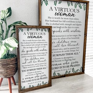 May include: Two rectangular wooden signs with dark brown frames featuring white backgrounds and black text. The text reads A VIRTUOUS WOMAN, followed by verses from Proverbs 31: 10-11; 25-30. Green leaf illustrations decorate the top and bottom edges.