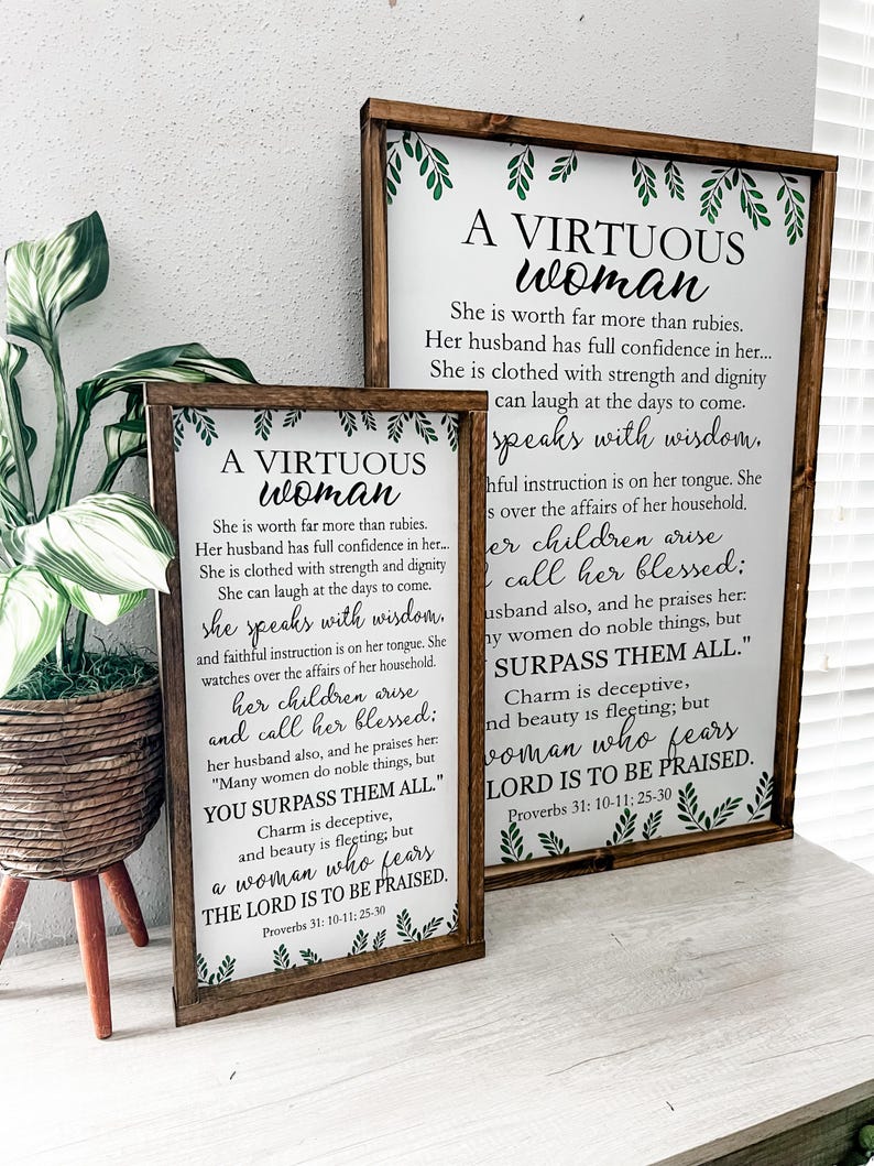 May include: Two rectangular wooden signs with dark brown frames featuring white backgrounds and black text. The text reads A VIRTUOUS WOMAN, followed by verses from Proverbs 31: 10-11; 25-30. Green leaf illustrations decorate the top and bottom edges.
