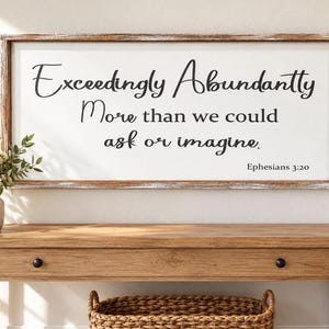 May include: A rectangular wall sign with a distressed brown wooden frame and a white background. The black text reads: Exceedingly Abundantly More than we could ask or imagine. Ephesians 3:20. The sign is mounted horizontally on a wall.