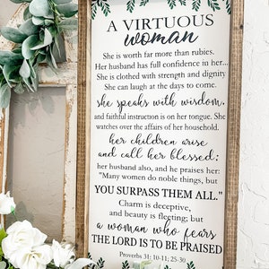 May include: A rectangular wooden sign with a dark brown frame and a white background featuring black text from Proverbs 31: 10-11, 25-30. The text reads A VIRTUOUS woman, She is worth far more than rubies, and includes green leaf accents at the top and bottom.