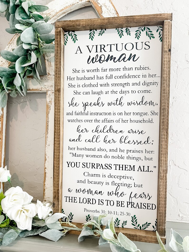 May include: A rectangular wooden sign with a dark brown frame and a white background featuring black text from Proverbs 31: 10-11, 25-30. The text reads A VIRTUOUS woman, She is worth far more than rubies, and includes green leaf accents at the top and bottom.