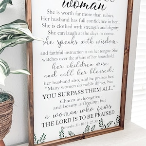 May include: A rectangular wooden framed sign with a white background featuring green leaf illustrations at the top and bottom. The text reads: A VIRTUOUS woman, She is worth far more than rubies. Her husband has full confidence in her... Proverbs 31: 10-11; 25-30.