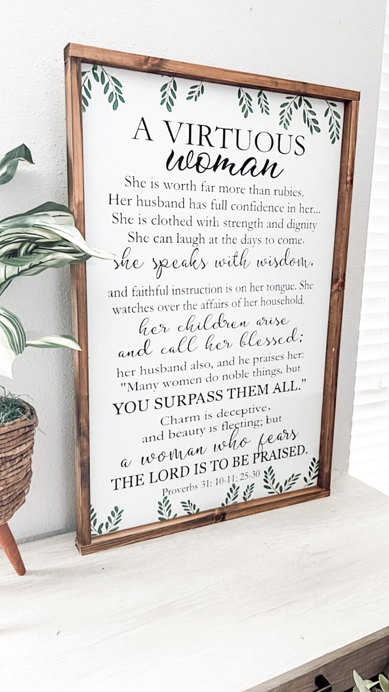 May include: A rectangular wooden framed sign with a white background featuring green leaf illustrations at the top and bottom. The text reads: A VIRTUOUS woman, She is worth far more than rubies. Her husband has full confidence in her... Proverbs 31: 10-11; 25-30.