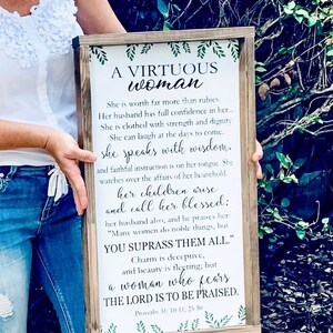 May include: A rectangular wooden framed sign with a white background featuring green leaf illustrations at the top and bottom. The text reads: A VIRTUOUS woman, She is worth far more than rubies. Her husband has full confidence in her... Proverbs 31: 10-11, 25-30.