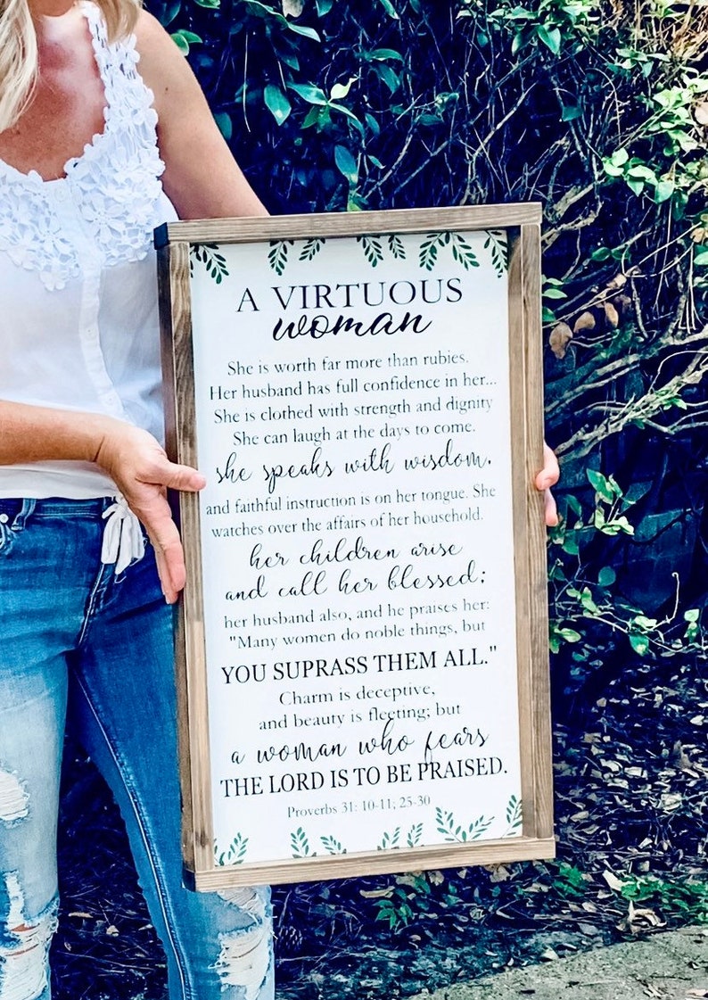May include: A rectangular wooden framed sign with a white background featuring green leaf illustrations at the top and bottom. The text reads: A VIRTUOUS woman, She is worth far more than rubies. Her husband has full confidence in her... Proverbs 31: 10-11, 25-30.