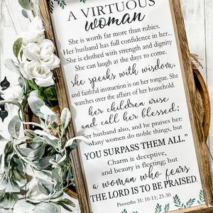May include: A rectangular wooden framed sign with a white background featuring green leaf illustrations at the top and bottom. The text reads: A VIRTUOUS WOMAN, She is worth far more than rubies, Proverbs 31: 10-11; 25-30.