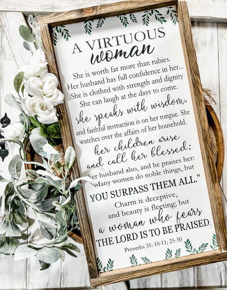 May include: A rectangular wooden framed sign with a white background featuring green leaf illustrations at the top and bottom. The text reads: A VIRTUOUS WOMAN, She is worth far more than rubies, Proverbs 31: 10-11; 25-30.