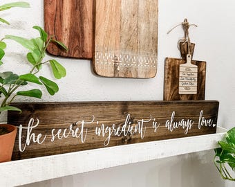 May include: A dark brown wooden sign with white cursive text that reads, the secret ingredient is always love. The sign is positioned horizontally on a white shelf, with two large wooden cutting boards and a small decorative board hanging above.