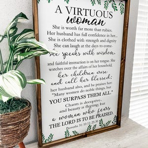 May include: A rectangular wooden sign with a dark brown frame featuring white background and black text that reads A VIRTUOUS WOMAN, followed by verses from Proverbs 31: 10-11; 25-30. The sign is decorated with green leaf illustrations at the top and bottom.