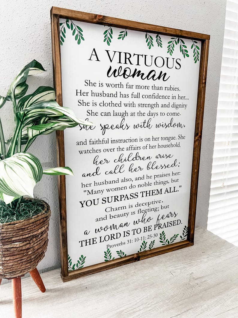 May include: A rectangular wooden sign with a dark brown frame featuring white background and black text that reads A VIRTUOUS WOMAN, followed by verses from Proverbs 31: 10-11; 25-30. The sign is decorated with green leaf illustrations at the top and bottom.