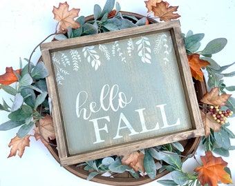 May include: A square wooden sign with the words "hello FALL" in white script. The sign is surrounded by a brown wooden frame and decorated with white leaf designs. The sign is placed on a woven basket with fall foliage.