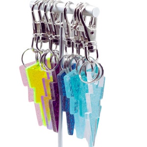 May include: A collection of lightning bolt shaped keychains hanging from a white display stand. Each keychain features a glittery finish in various colors including neon yellow, purple, blue, and white, attached to a silver metal lobster clasp.
