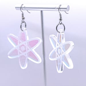 May include: A pair of iridescent atom earrings. The earrings are made of clear acrylic with a pink and blue iridescent finish. The atom symbol is outlined in silver.