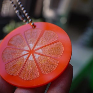 May include: Orange slice keychain made of bright orange plastic with a silver chain. The keychain is shaped like a slice of orange with a textured surface.