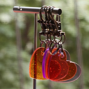 May include: A set of heart-shaped keychains in shades of orange, red, and purple, hanging from a silver metal display stand. The keychains have glitter accents and are attached to silver clasps.