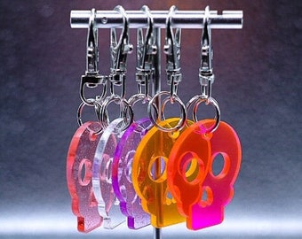 May include: Five translucent acrylic skull keychains in red, clear, purple, yellow, and pink, each featuring a glittery finish and attached to a silver-toned metal swivel lobster clasp and split ring, hanging from a horizontal metal display bar.