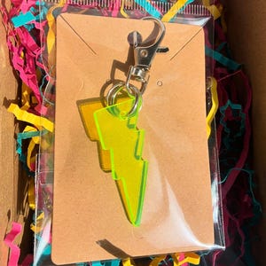May include: A neon yellow lightning bolt keychain made of translucent acrylic material. The charm is attached to a silver-toned metal lobster claw clasp and a split ring, mounted on a rectangular brown cardboard display card inside a clear plastic bag.