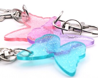 May include: Three butterfly-shaped keychains made of translucent, glitter-infused resin in shades of pink, purple, and blue. Each butterfly is attached to a silver-toned metal split ring and a lobster claw clasp for secure attachment to items.
