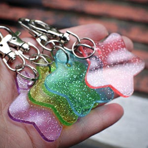 May include: A set of four star-shaped keychains made of translucent, glitter-infused acrylic in shades of purple, yellow, green, and pink. Each star is attached to a silver-toned metal lobster clasp by a small circular jump ring.