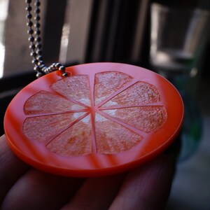 May include: A red and orange plastic pendant shaped like a slice of citrus fruit with a white center. The pendant is on a silver chain.