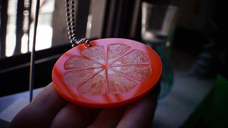 May include: A red and orange plastic pendant shaped like a slice of citrus fruit with a white center. The pendant is on a silver chain.