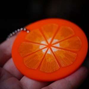 May include: A bright orange keychain shaped like a slice of orange. The orange slice has a white center and is detailed with lines to resemble the segments of an orange.